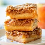 A stack of 3 apple pie bars on a white plate with a red apple in the background off to the right.