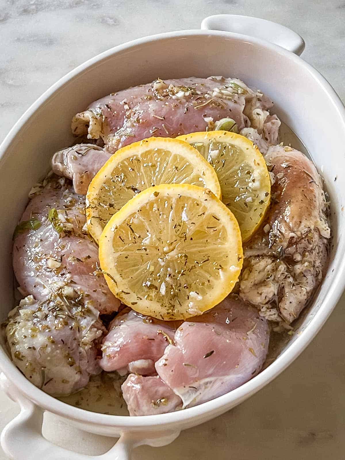 Marinated chicken thighs with lemon slices in a white dish ready for the oven.