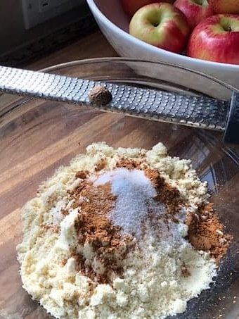 Dry ingredients, unmixed in a clear glass bowl with micro plane and whole nutmeg balanced on side. White bowl of Honeycrisp apples in upper right corner of image.