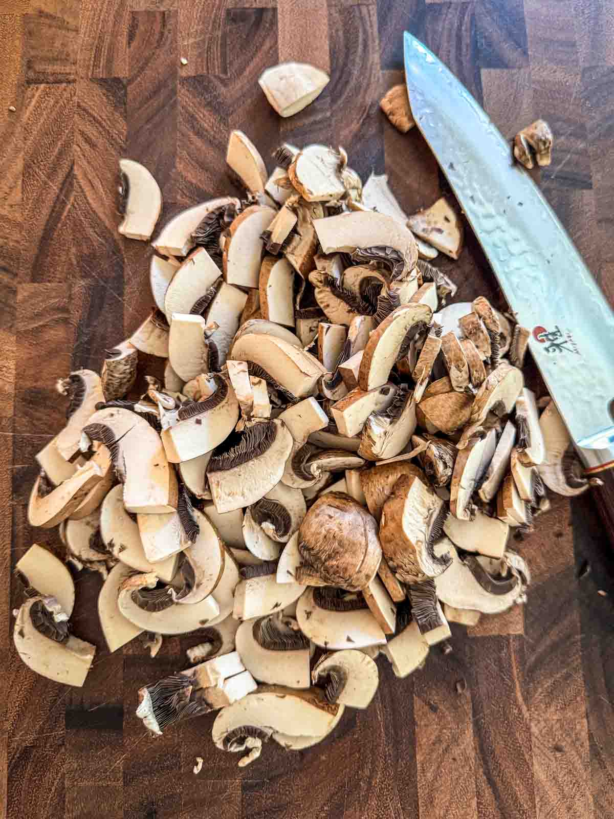 Thinly sliced Crimini mushrooms on a cutting board with a chef's knife.
