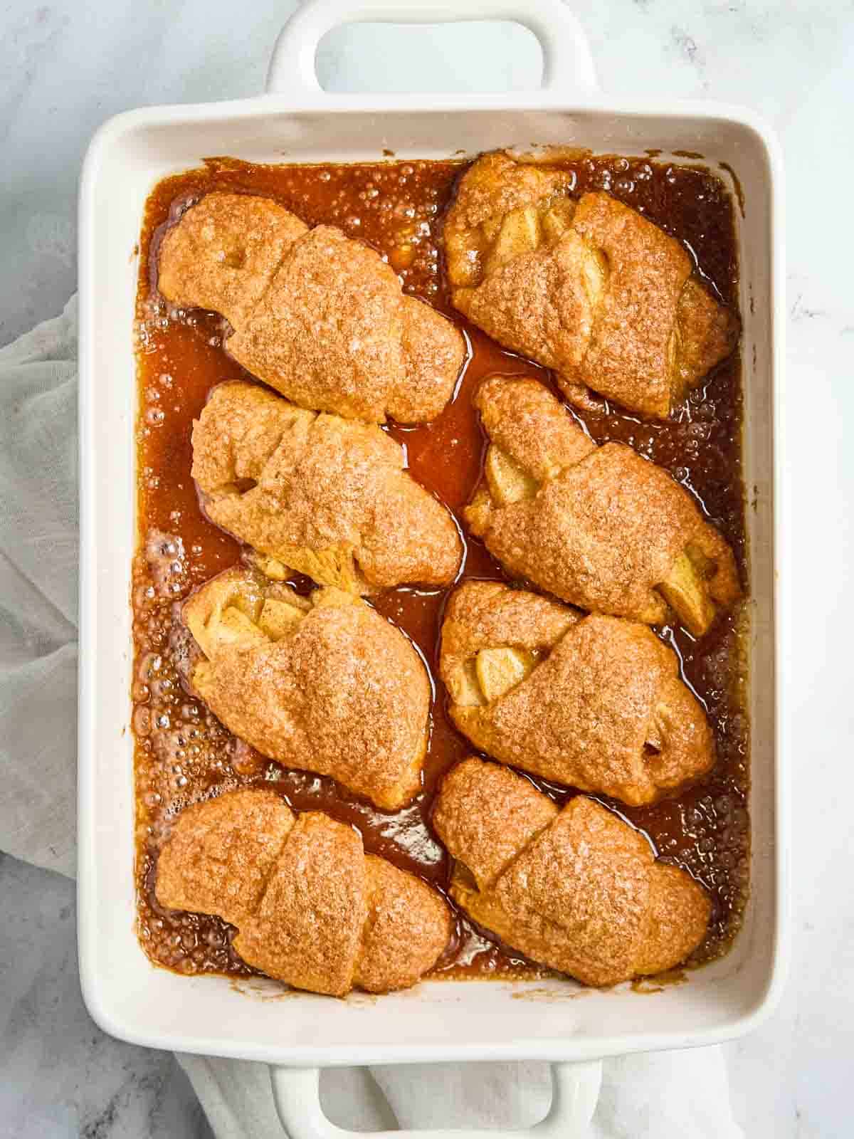 Golden brown crescent roll apple dumplings baked in a white dish, bubbling with cinnamon sugar glaze.
