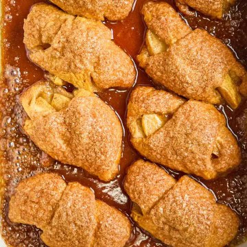 Golden brown crescent roll apple dumplings baked in a white dish, bubbling with cinnamon sugar glaze.