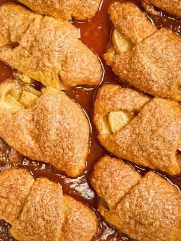 Golden brown crescent roll apple dumplings baked in a white dish, bubbling with cinnamon sugar glaze.