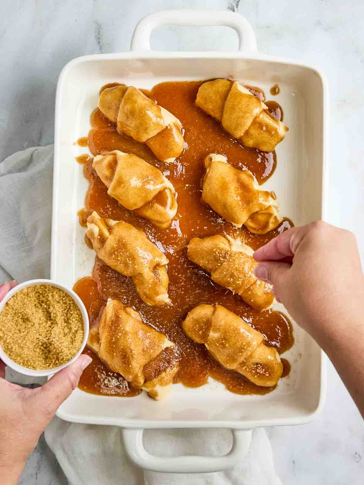 A hand sprinkling sugar on crescent roll apple dumplings in a white dish, with cinnamon sugar glaze reeady for the oven.