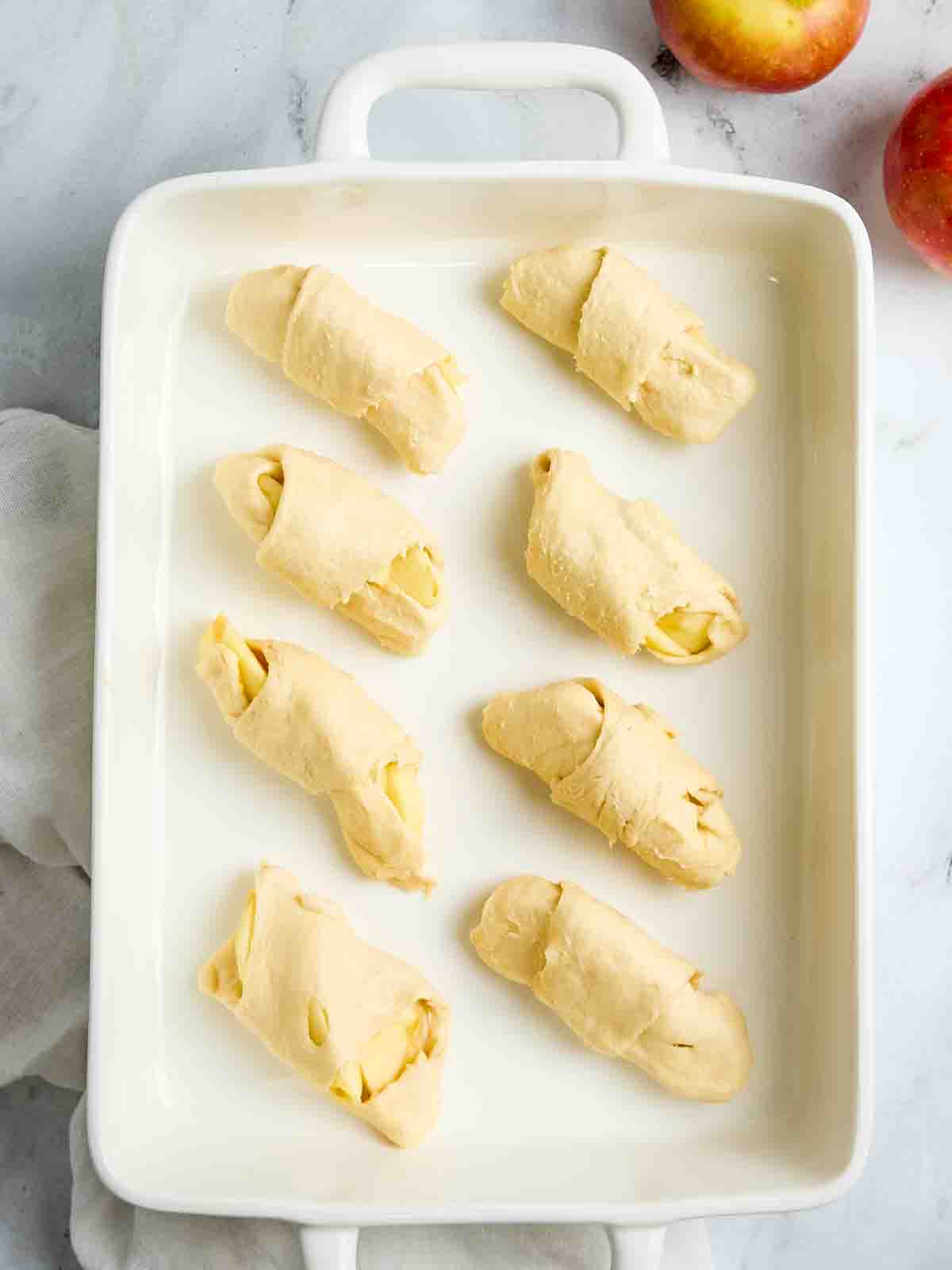 Crescent rolls rolled with sliced apples in a white baking dish for crescent roll apple dumplings recipe.