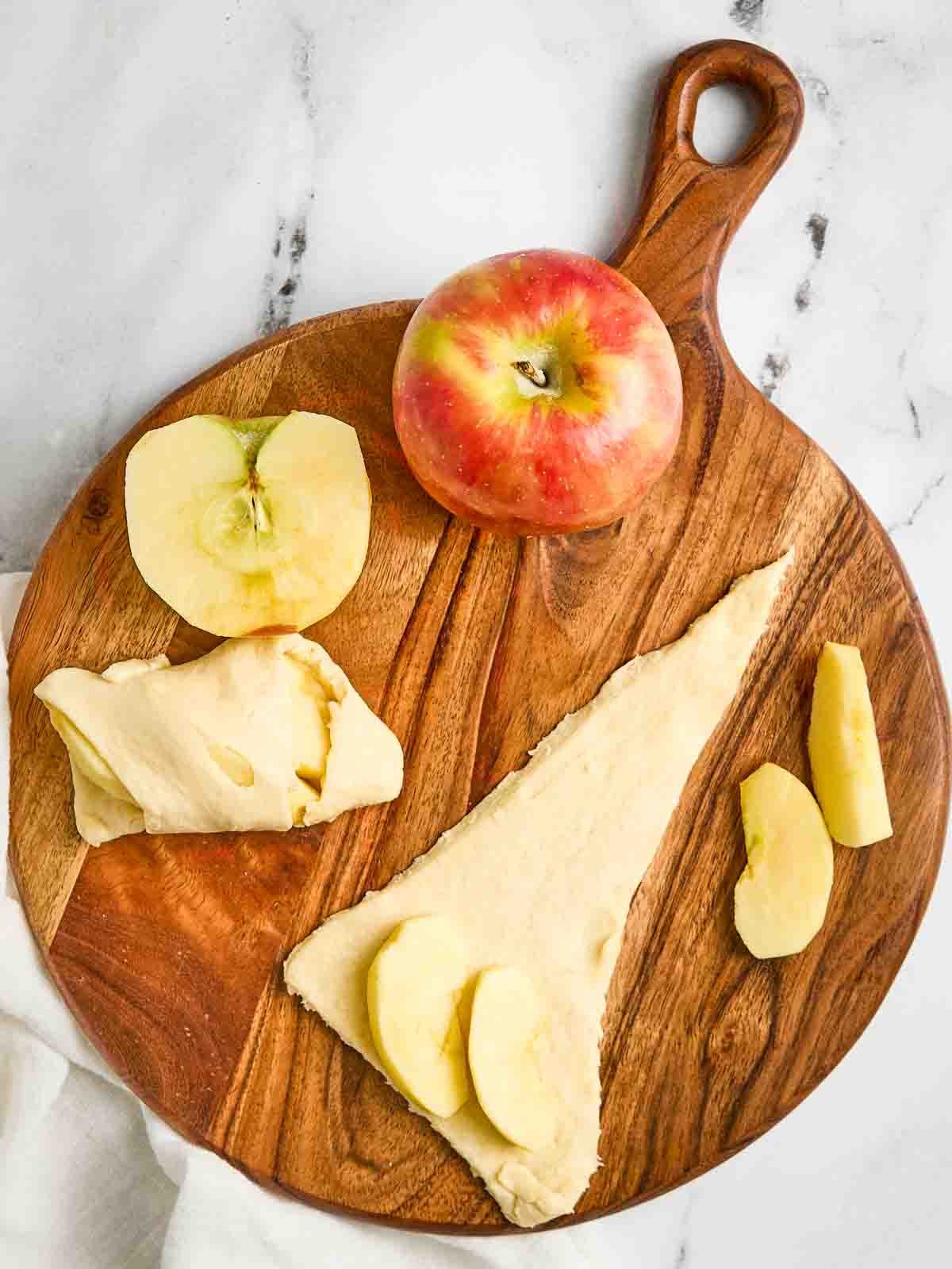 Crescent roll triangle with two thin apple slices on the wide end, one rolled crescent, and a whole Honeycrisp apple on a wooden cutting board.