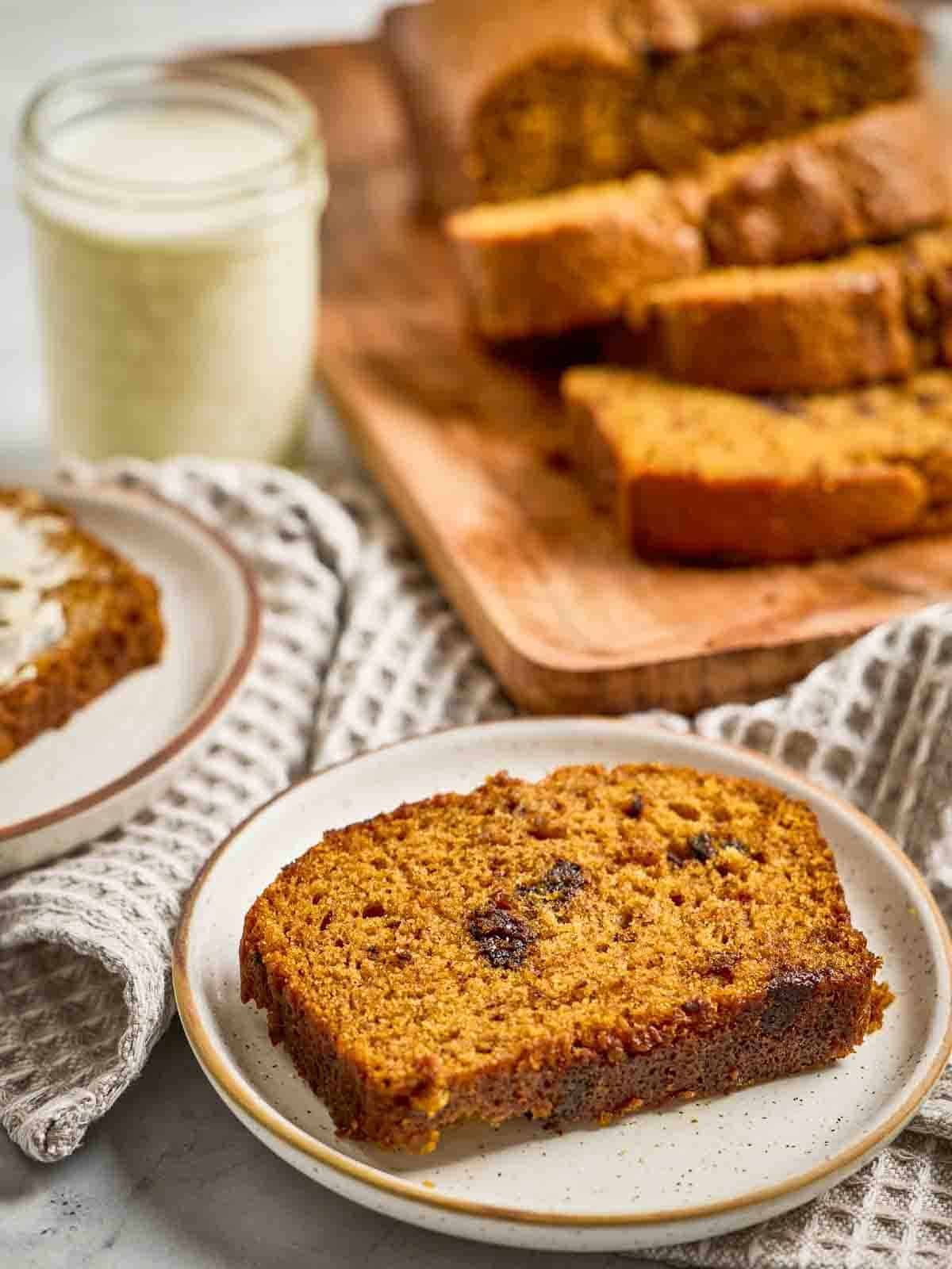 A slice of pumpkin chocolate chip bread on a white plate with sliced loaf in background, o checkered napkin.