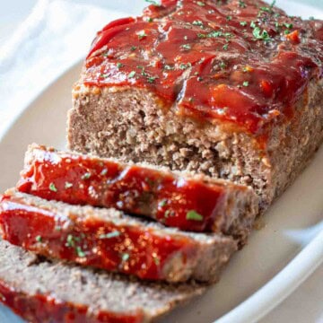 The best meatloaf with ketchup topping sliced on a white platter and garnished with chopped parsley.