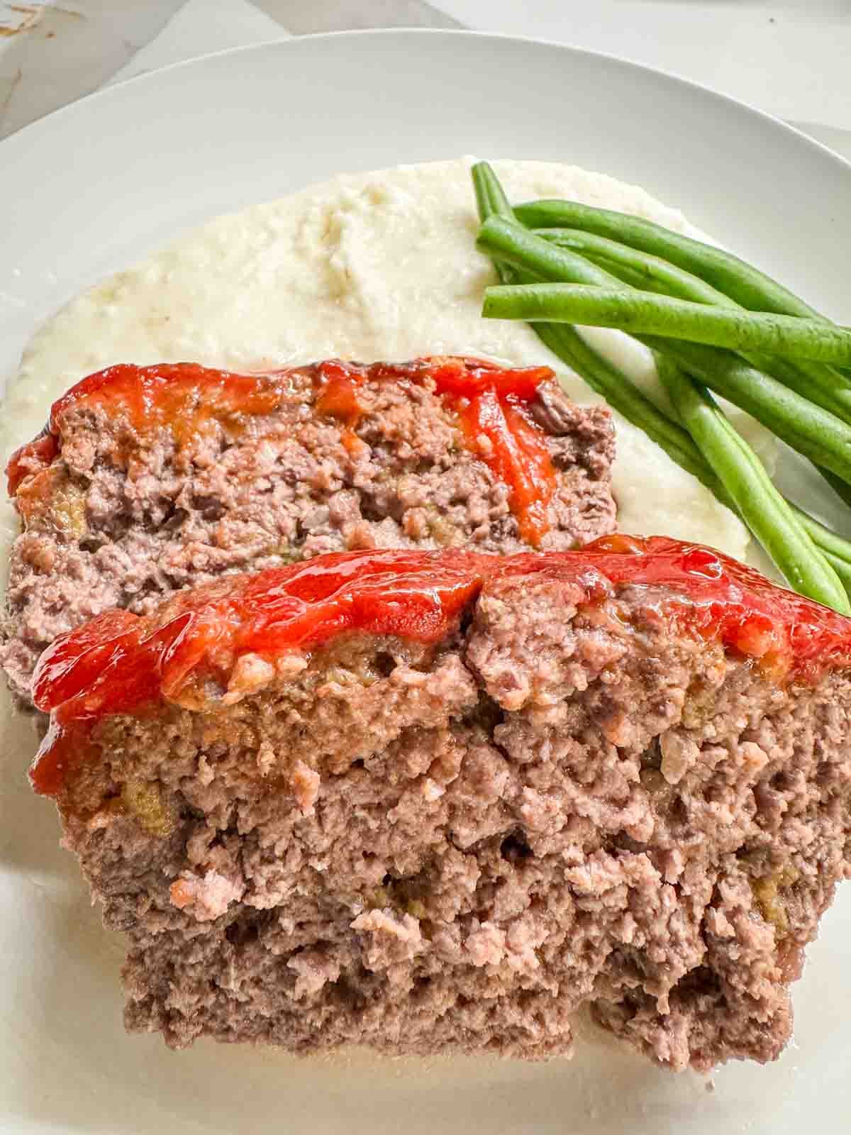 Two slices of tender & juicy meatloaf over mashed potatoes with a side of green beans.