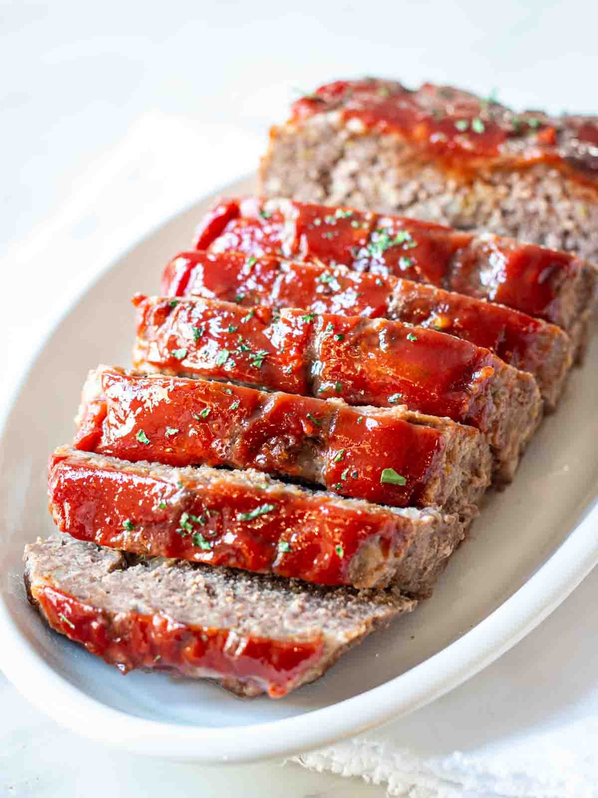 Classic meatloaf with ketchup topping sliced on a white platter and garnished with chopped parsley.