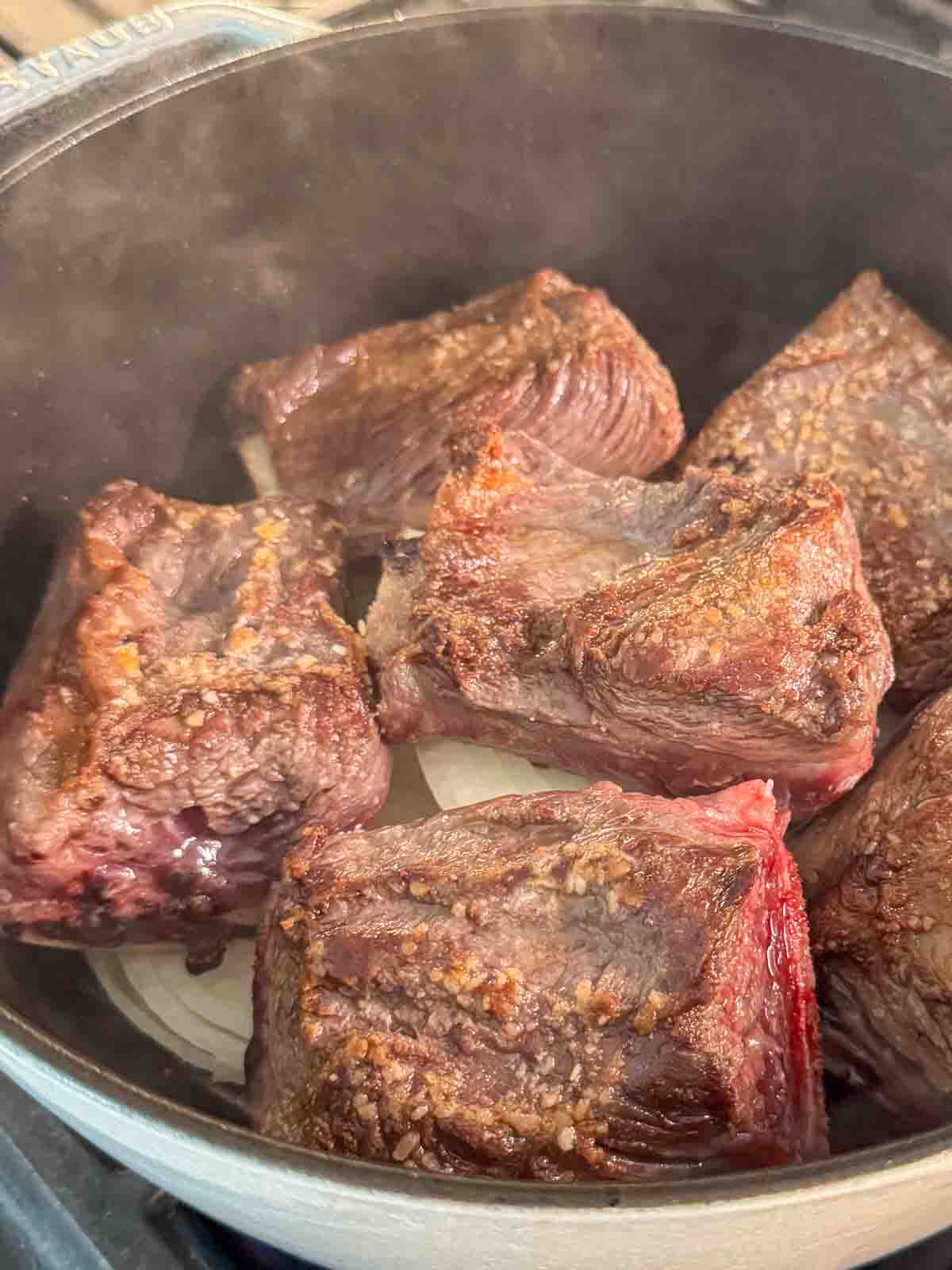Seared beef short ribs layered on top of thick round slices of onion inside a Dutch oven.