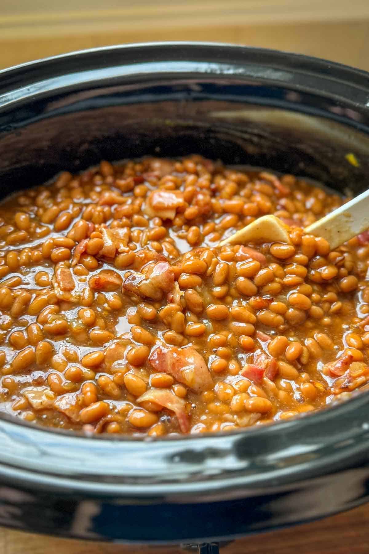 Close-up of homemade baked beans in a slow cooker, showing a rich, thick, and hearty bean mixture with a spoon for serving.